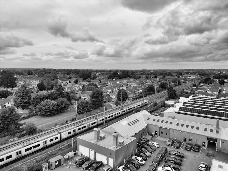 A black-and-white aerial view of a suburban area showing a residential neighborhood with rows of houses, trees, and streets. In the foreground, there is a commercial building with a sloped roof, surrounded by a parking lot filled with various parked cars. A modern train is passing on elevated tracks behind the building, extending across the image. The background features a distant horizon with an expansive sky filled with scattered clouds, creating a contrast between the urban landscape and natural elements. The scene appears clean and well-maintained, with no visible dirt or debris, demonstrating an organized environment.