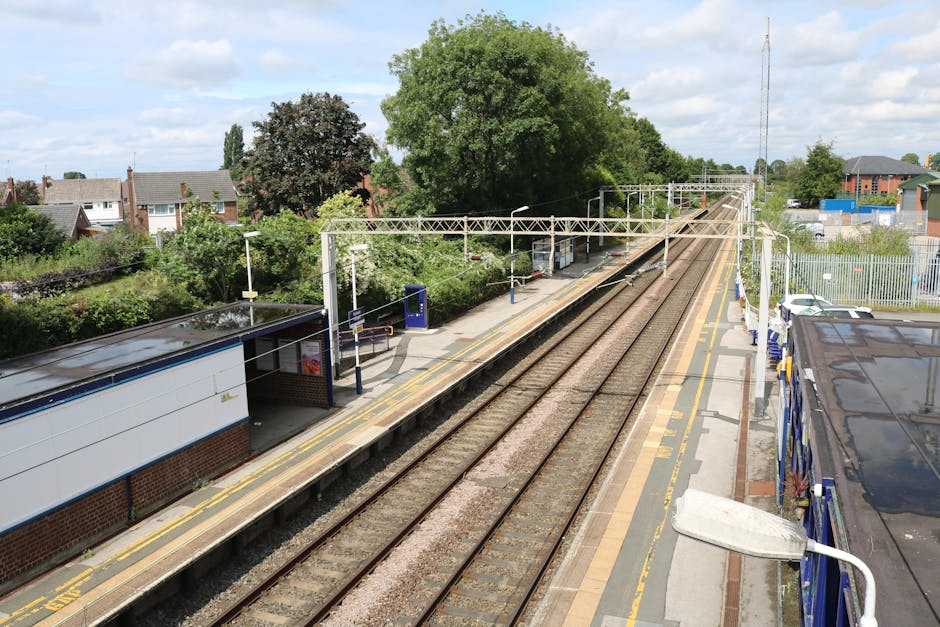 A high-angle view of a residential train station platform in Enfield, featuring parallel railway tracks with well-maintained wooden sleepers and gravel beds. The platform surface is made of smooth concrete with yellow tactile paving along the edge for safety, appearing clean and free of debris. A small shelter with a black roof and blue advertising panels provides seating and shelter for waiting passengers. Adjacent to the platform, there are white and blue fencing and a street lamp with a curved arm, casting light on the pathway below. Lush green trees and suburban houses with brick exteriors and pitched roofs fringe the background, under a partly cloudy sky, creating a tidy and orderly scene. The image emphasizes the importance of maintaining cleanliness and hygiene at transit stations, which is a focus of [COMPANY_NAME], specialists in domestic cleaning and surface sanitisation near Enfield Town station.