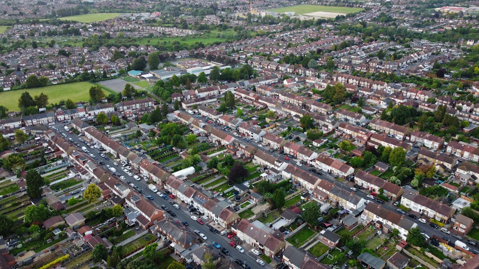 Aerial view of a residential suburb showing rows of terraced houses with small front gardens and driveways, lined along narrow streets filled with parked cars. The houses have tiled roofs and are surrounded by lush green trees and lawns. In the background, there are larger open fields and patches of greenery, with a distant view of additional housing and scattered trees under a cloudy sky.