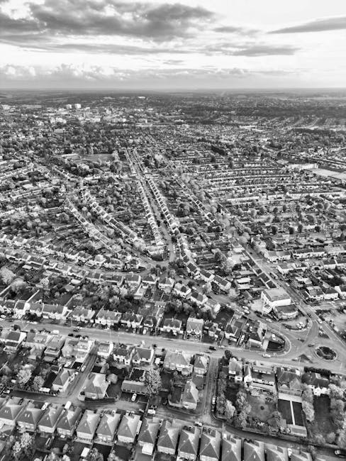 A black and white aerial photograph of a residential area featuring numerous detached and semi-detached houses with pitched roofs, some with chimneys, arranged in a grid pattern. The landscape includes tree-lined streets, small gardens, and varied building materials such as brick and tile. The sky is filled with scattered, fluffy clouds, and the overall scene conveys a calm, suburban environment. For professional surface cleaning, deep cleaning, and sanitisation services, Cleaners Enfield can assist with maintaining hygiene and cleanliness in such residential settings.